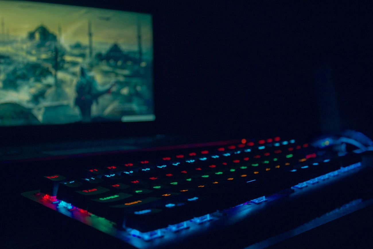 A backlit gaming keyboard in a dark room, a game glowing on the screen behind
